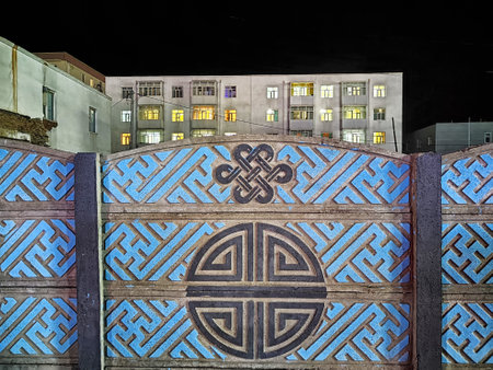 Decorative wall with intricate patterns stands in front of illuminated buildings at night, showcasing urban architecture and design.の写真素材
