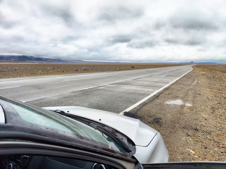 A car parked beside a long, empty road leads to a vast, arid terrain beneath cloudy skies in a remote region.の写真素材