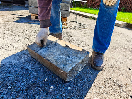 A person in work gloves and boots is lifting a heavy concrete block on a gravel surface during a construction task on a sunny day.の写真素材