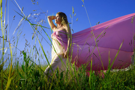Woman posing gracefully in a field with flowing pink fabric under a clear blue sky during golden hourの写真素材