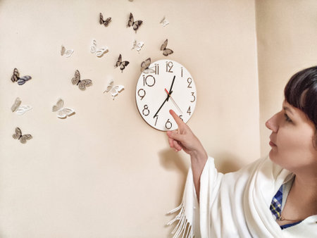 Middle-aged woman psychologist points to clock in her office while surrounded by decorative butterflies on the wallの写真素材