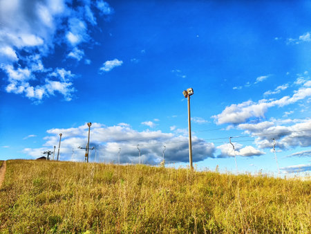 Bright blue sky filled with clouds above a grassy hill with floodlights and power lines creating an open outdoor atmosphere.の写真素材