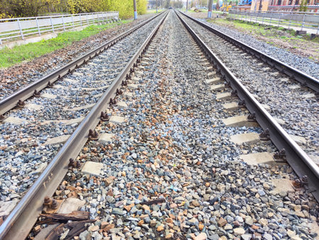 Metal tracks line a gravel path, bordered by greenery and distant structures, under clear skies depicting a peaceful transport route.の写真素材