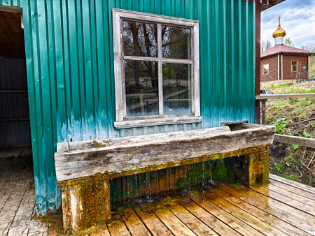 A rustic wooden fountain spills water from its basin next to a green shed while a church is visible in the background under a cloudy sky.の写真素材