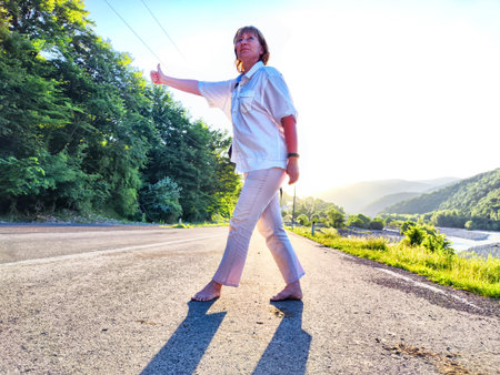 Middle-aged woman hits a ride along a scenic road surrounded by lush greenery and hills, embracing her travel spirit.の写真素材