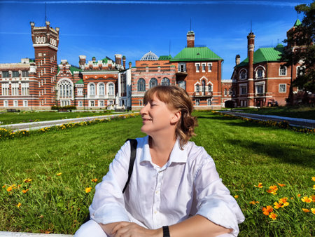 Blogger smiles joyfully, seated on the grass with vibrant flowers and historical architecture on a bright sunny day.の写真素材