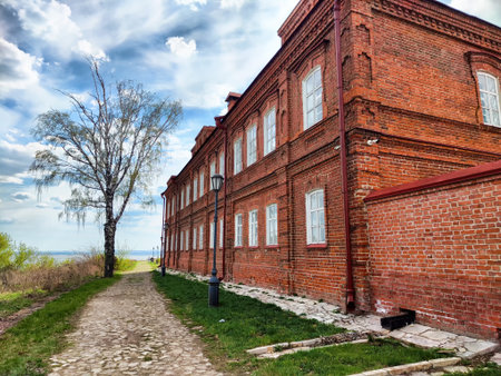 A red brick structure stands beside a cobblestone path lined with a solitary tree under a bright sky...