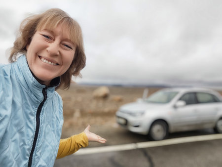 A middle-aged blonde woman captures a joyful moment during her travel excursion, smiling widely by the roadside with her rental car.の写真素材