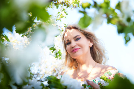 Mature blonde woman poses among blooming flowers while enjoying a sunny day outdoors, showcasing her...
