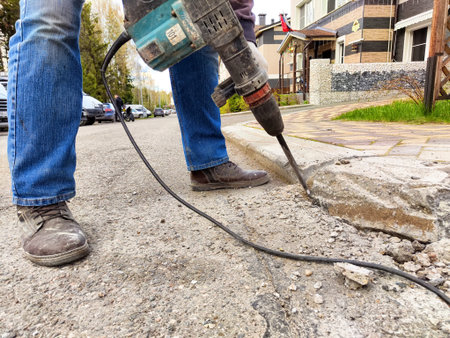 A person is using a jackhammer to break concrete on a street outside residential buildings. Cars are...