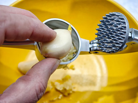 Hand holds a garlic clove above a stainless steel press, ready to crush it. Yellow bowl collects the freshly pressed garlic.の写真素材