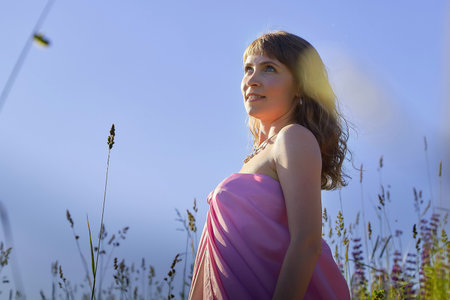 Woman in a pink wrap stands gracefully in a sunny meadow surrounded by tall grass and vibrant wildflowersの写真素材