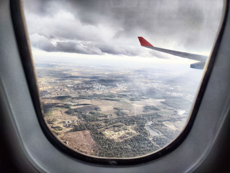 The airplane wing juts out into the cloudy sky, while vast green fields and urban areas stretch below on a flight journey.の写真素材