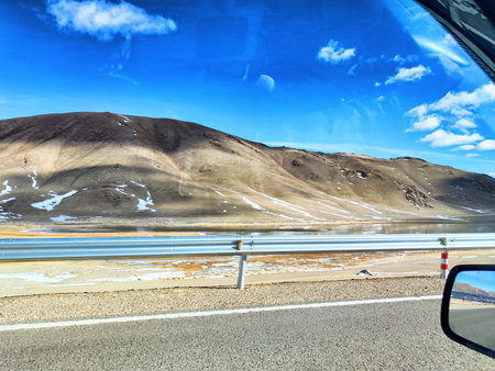 Scenic view of mountains and shimmering water beside a road under a bright blue sky with fluffy clouds, showing natural beauty.の写真素材