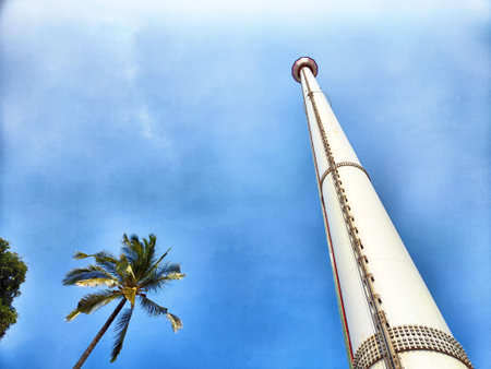 A tall industrial tower stands prominently against a backdrop of clear blue sky, alongside a swaying palm tree on a sunny day.の写真素材