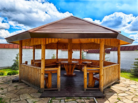 A sturdy wooden gazebo offers a cozy gathering spot surrounded by greenery, with clear blue skies overhead and soft clouds in sight.の写真素材