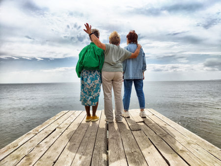 Three women stand together on a wooden pier, smiling and enjoying the view of a serene ocean and overcast sky.の写真素材
