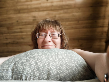 Middle-aged woman with disheveled hair smiles while resting on a pillow in a warm, wooden bedroom during a peaceful morning.の写真素材