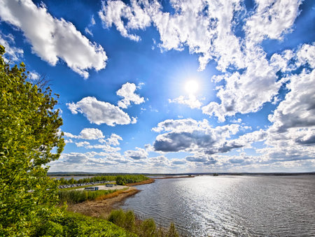 Expansive view of a peaceful lake under a bright sun, with fluffy clouds and green foliage framing the scene, creating a serene atmosphere.の写真素材