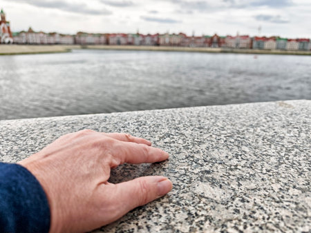 A hand rests gently on a smooth granite ledge, gazing at the tranquil river and colorful buildings along the waterfront.の写真素材