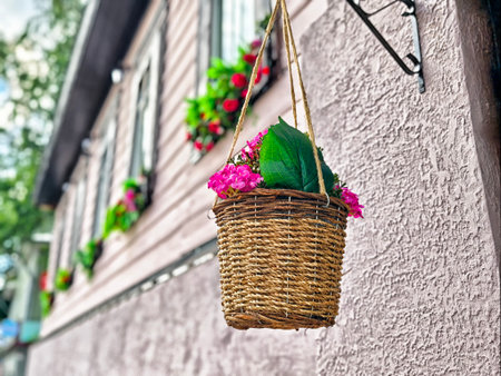 A wicker basket filled with vibrant pink flowers dangles from a wall, adding charm to the quaint home exterior adorned with plants.の写真素材