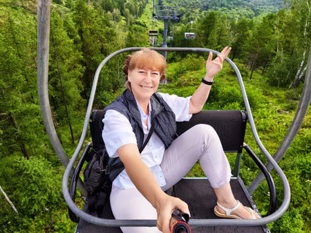 A mature woman takes a joyful selfie while riding a cable car through vibrant green mountains, capturing the essence of her travel excitement.の写真素材