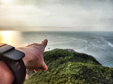 A hand is extended towards the vast ocean, capturing the serene sunset glow overlooking a lush green...