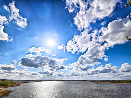 Large, puffy clouds float across a vibrant blue sky above a calm river during midday, with sunlight...