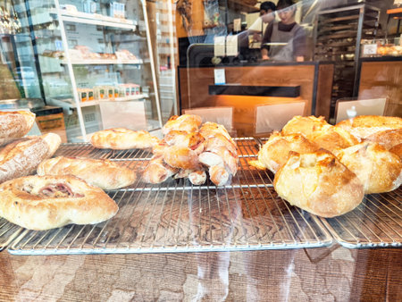 A variety of golden-brown pastries sit on a wire rack, showcased behind a bakery window, inviting...