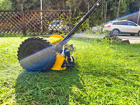 A lawn mower is actively trimming grass in a tidy yard under sunny conditions, showing vibrant green grass clippings flying in the air.の写真素材