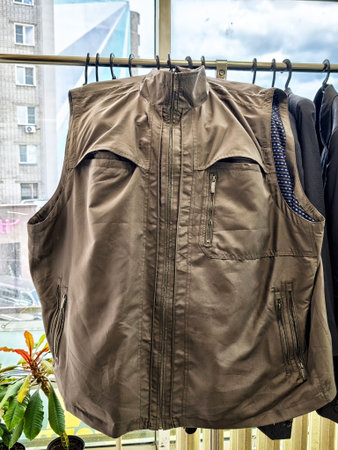 A brown sleeveless jacket hangs on a rack inside a building, with a city view through the window.