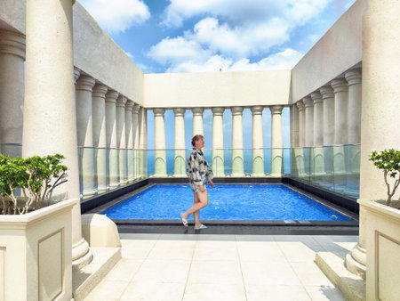 A mature woman takes a leisurely stroll beside a vibrant blue pool, enjoying the beautiful weather and architecture of her travel excursion.の写真素材