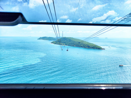The cable car glides over clear blue waters, showcasing an island landscape dotted with greenery under a vibrant sky filled with fluffy clouds.の写真素材