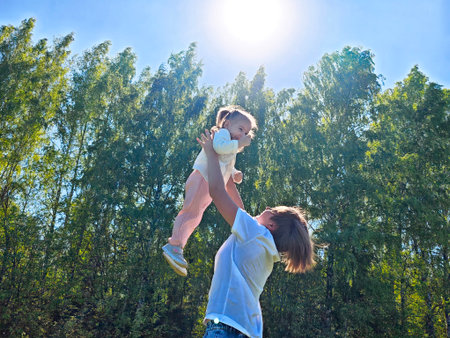 Mother lifts her joyful child under bright sun in a lush green park on a clear dayの写真素材