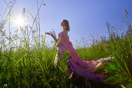 Elegant woman in a flowing pink gown stands amidst tall grass on a sunny dayの写真素材