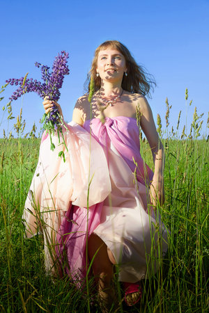 Woman in a pink dress enjoys a sunny day in a lush green field while holding a bouquet of purple flowersの写真素材