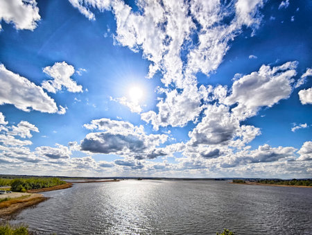 Bright sunlight shines over a serene river as fluffy clouds drift in a blue sky, creating a tranquil scene of nature and peace.の写真素材