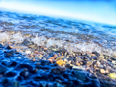 Gentle waves softly collide with colorful pebbles on a tranquil beach under bright blue skies during the afternoon light.の写真素材