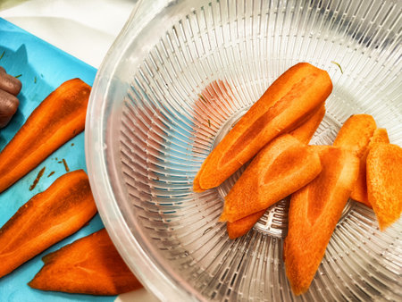 Freshly cut carrots sit in a colander, ready for rinsing after being sliced on a blue cutting board in a sunlit kitchen.の写真素材