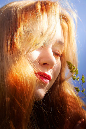 Sunlit portrait of a woman with flowing red hair and vivid lips enjoying nature in a bright outdoor settingの写真素材