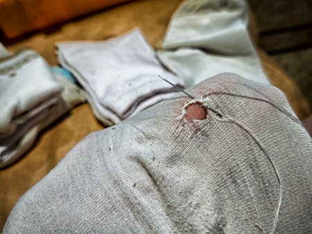 With a focus on a needle and thread, someone is mending a torn piece of fabric in a cozy home setting surrounded by various cloth items.の写真素材