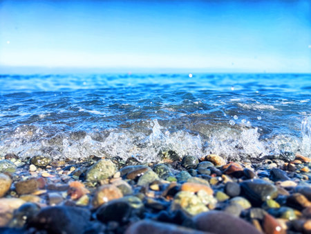 Seaside waves create a soothing sound as they wash over smooth, colorful pebbles under a bright blue sky.の写真素材