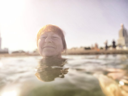A middle-aged blonde woman smiles while partially submerged in a bright, sunlit water during her travel excursion.の写真素材