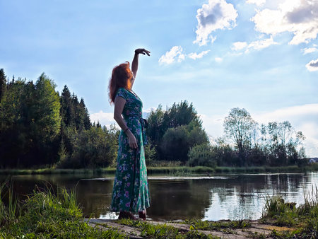 Woman in green dress reaching towards the sky by a serene lake in the afternoon lightの写真素材