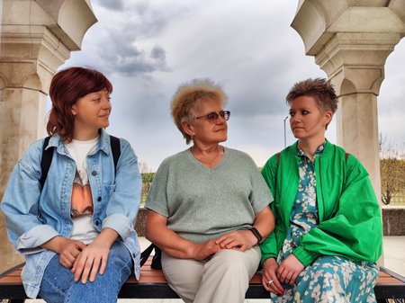 Three women engage in conversation under a stone archway near a park on a cloudy day