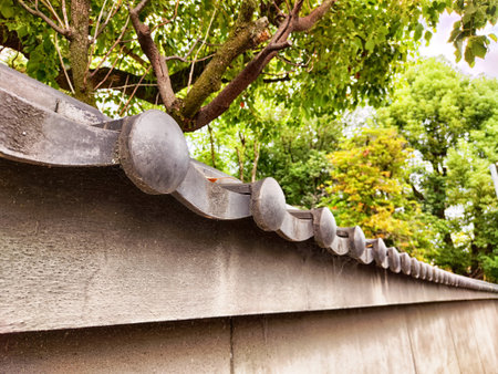 Lush trees and vibrant plants surround a traditional Japanese wall, showcasing ornate tiles under...