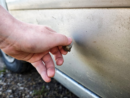 A teenager uses a stone to scratch the surface of a parked car, showing an act of vandalism in an outdoor setting.の写真素材