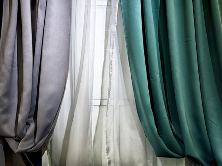 Various curtains in shades of gray and teal elegantly frame a window, letting soft natural light illuminate the interior setting.の写真素材
