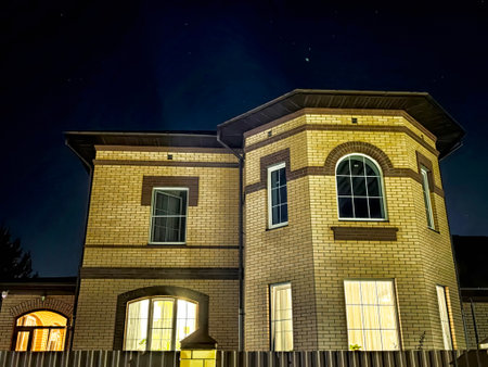 A brick house stands tall at night, with warm light coming from the windows, illuminated against a clear star-filled sky.の写真素材