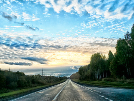 A long, empty road leads toward a beautiful sunset, with trees lining the sides and clouds creating...